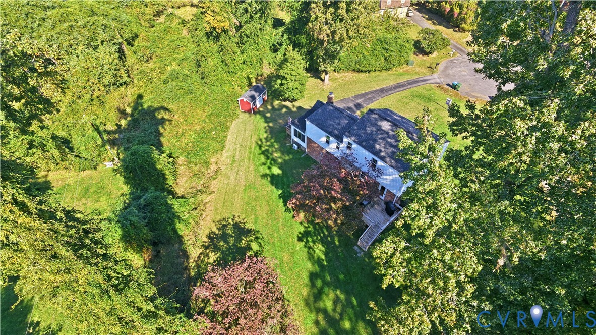 Aerial perspective of a multi-story residence featuring an attached wooden deck, paved driveway, detached storage structure, and expansive grounds with mature trees