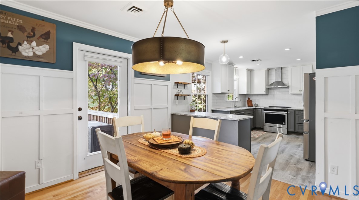Dining area featuring white board-and-batten wainscoting, an industrial-style drum pendant light, and wood-finish flooring, opening to a kitchen equipped with two-tone cabinetry and stainless steel appliances