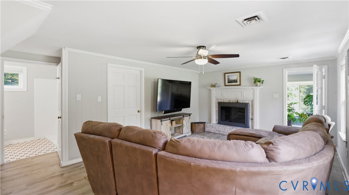 Living area featuring a painted brick fireplace with a decorative mantel, ceiling fan with integrated lighting, wood-finish flooring, and crown molding