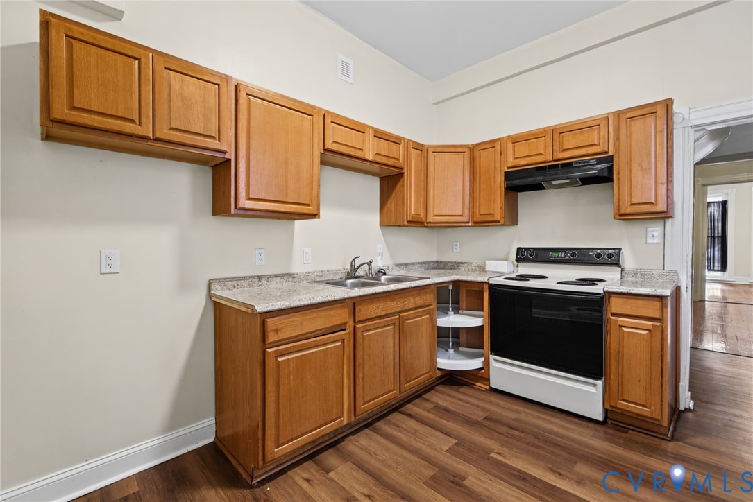 Kitchen featuring natural wood cabinetry, stone-patterned laminate countertops, wood-finish flooring, a stainless steel dual-basin sink, and an integrated corner carousel cabinet
