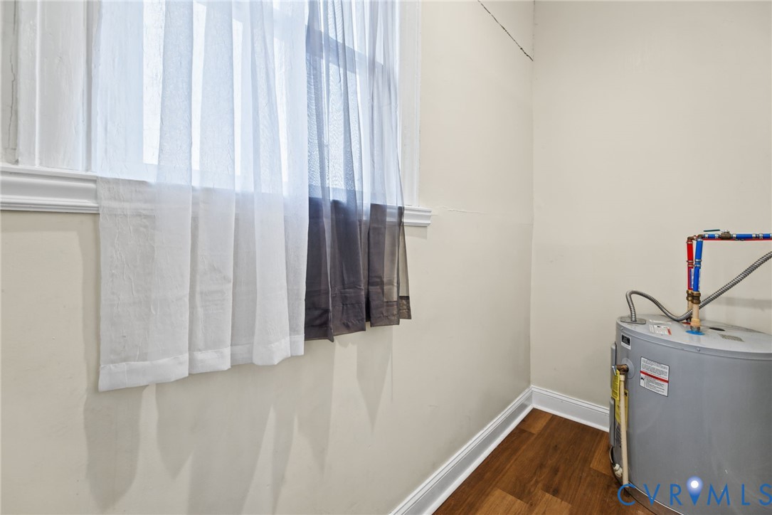 Electric water heater with exposed plumbing connections, wood-finish flooring, and single-pane window with white trim