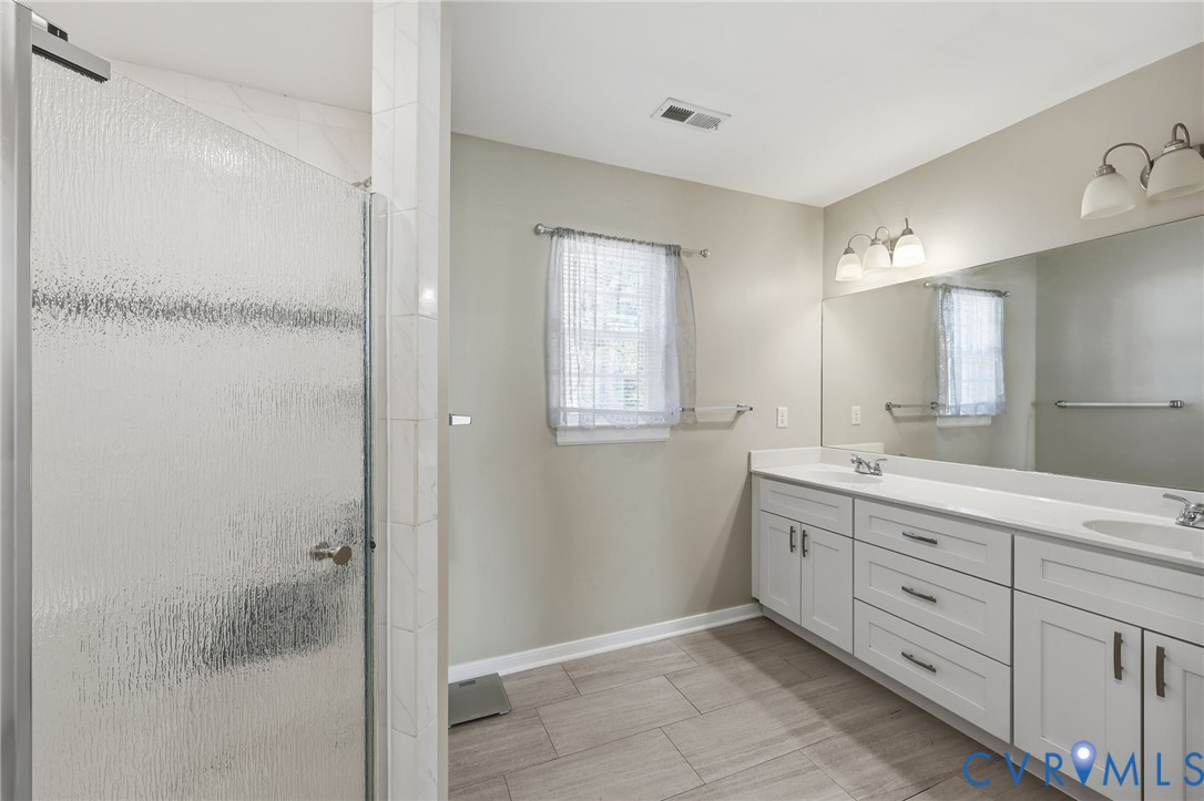 Dual-sink vanity with white cabinetry and solid surface countertop, walk-in shower with textured glass enclosure, ceramic tile flooring, and wall-mounted vanity lighting
