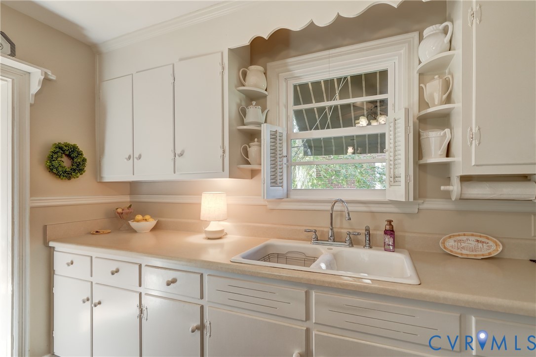 Kitchen featuring white cabinetry with integrated scalloped valance, solid-surface countertops, double-basin sink with metallic faucet, and decorative corner shelving