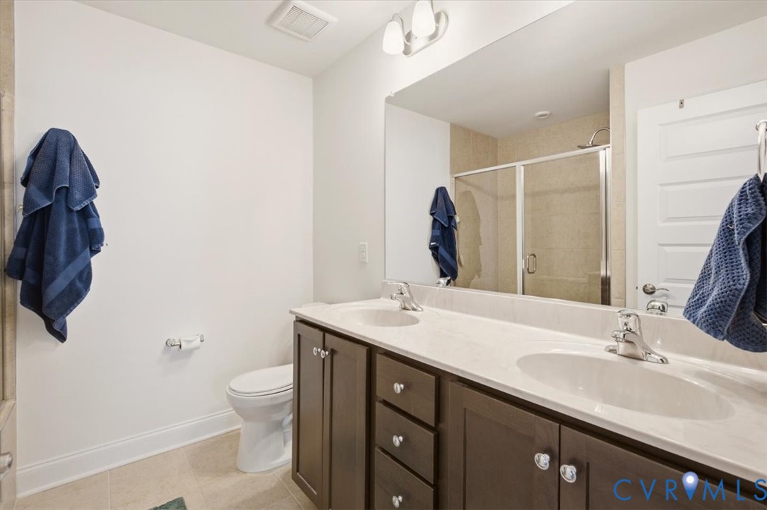 Dual-sink vanity with dark wood-finish cabinetry, solid surface countertop, large wall-mounted mirror, and glass-enclosed shower