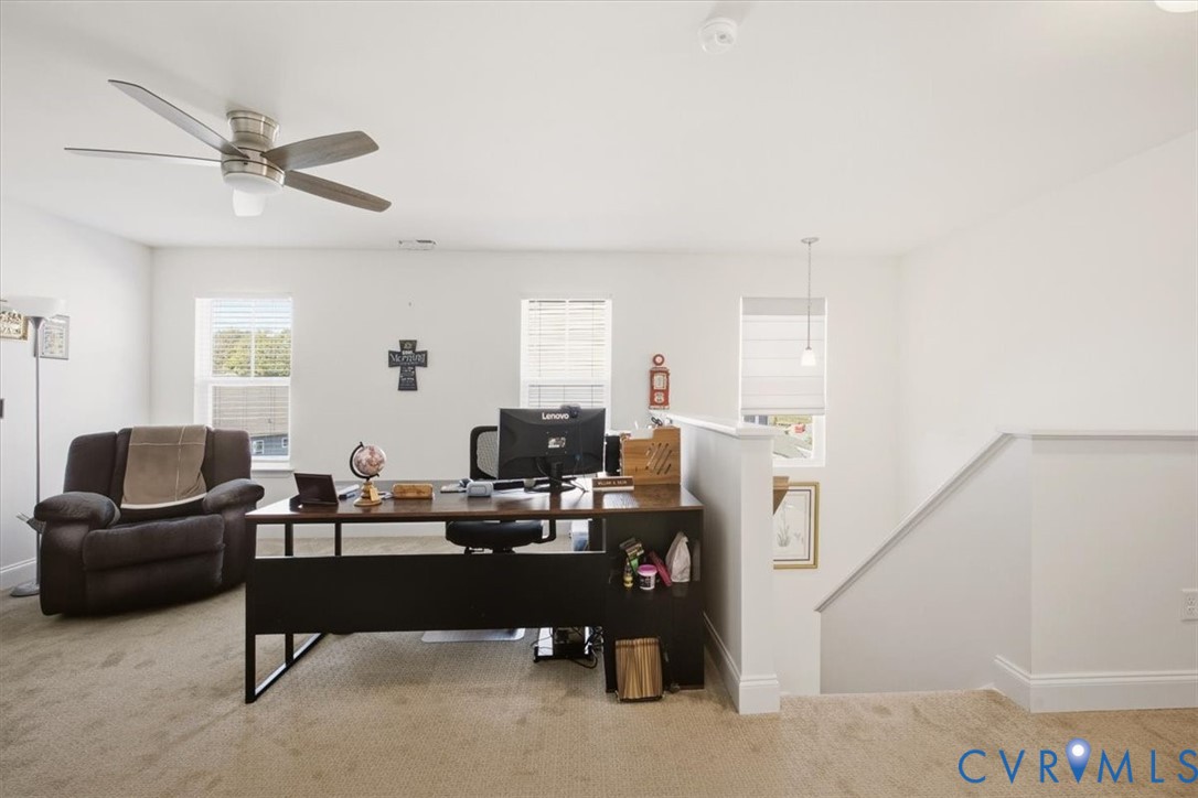 Open floor plan featuring carpeting, three windows with blinds, a ceiling fan with integrated lighting, and a half-wall stair railing