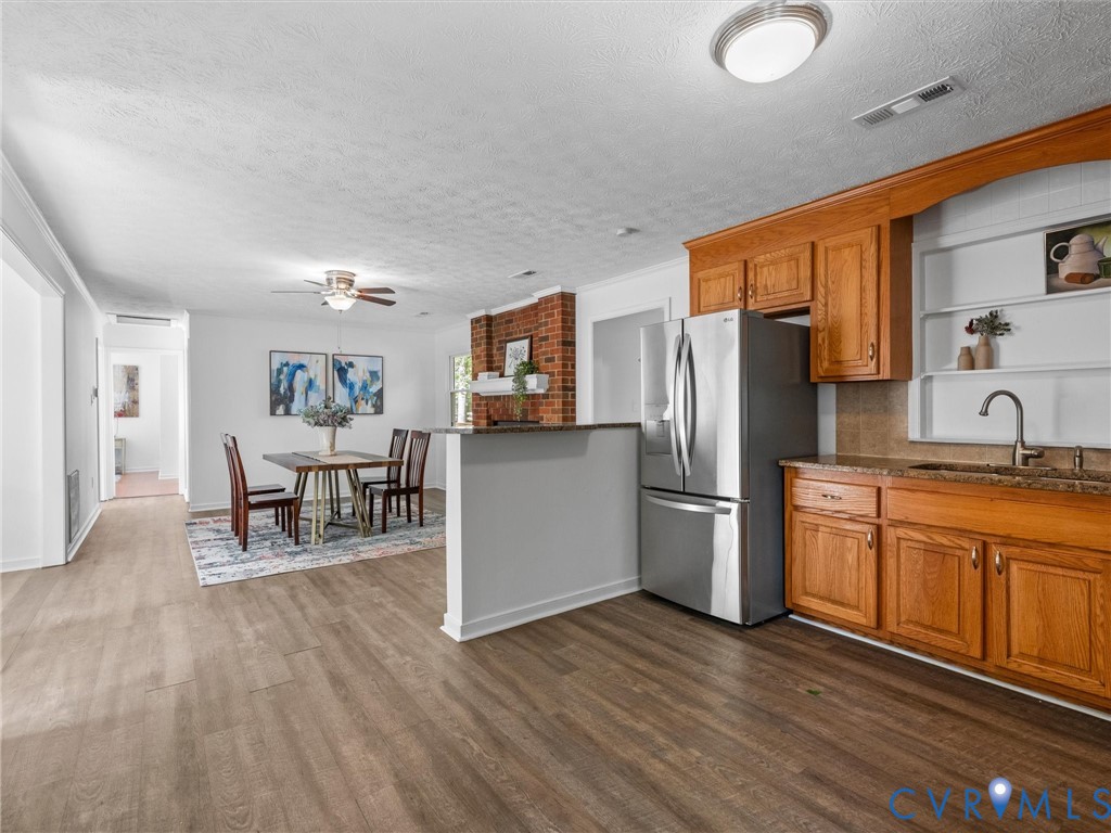 Kitchen featuring wood cabinetry, stone countertops, stainless steel refrigerator, and integrated shelving, complemented by wood-finish flooring and a red brick fireplace