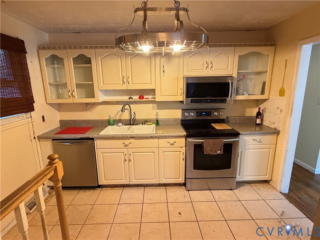 Kitchen featuring off-white cabinetry with glass-paneled uppers, stainless steel appliances, a double-basin sink, stone-finish countertops, and a decorative metal-frame overhead light fixture