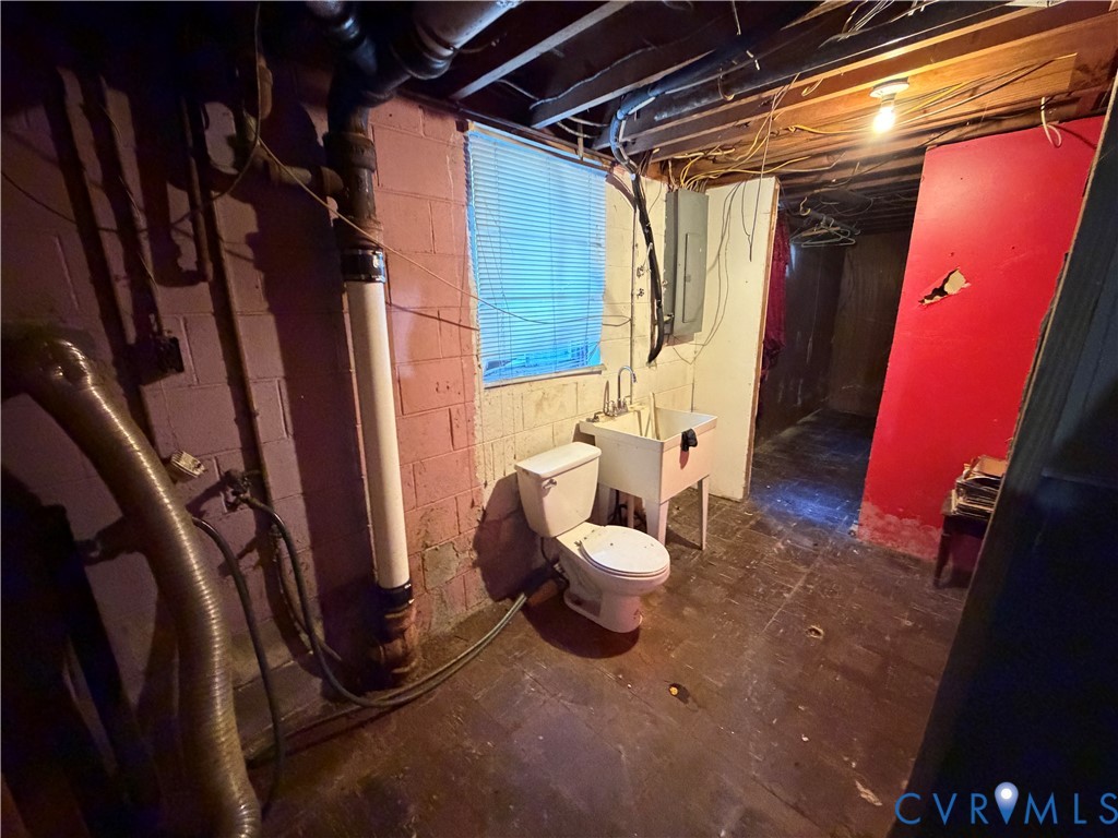 Utility space featuring concrete masonry walls, a floor-mounted toilet, a pedestal sink basin, and electrical panel