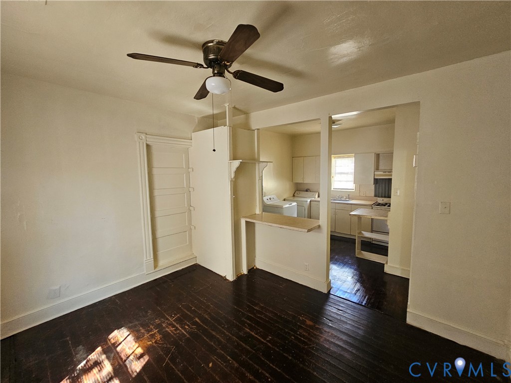 Dark-toned wood-finish flooring, central ceiling fan with integrated light fixture, built-in shelving, and a peninsula counter separating the kitchen area Dark-toned wood-finish flooring, central ceiling fan with integrated light fixture, built-in shelving, and a peninsula counter separating the kitchen area