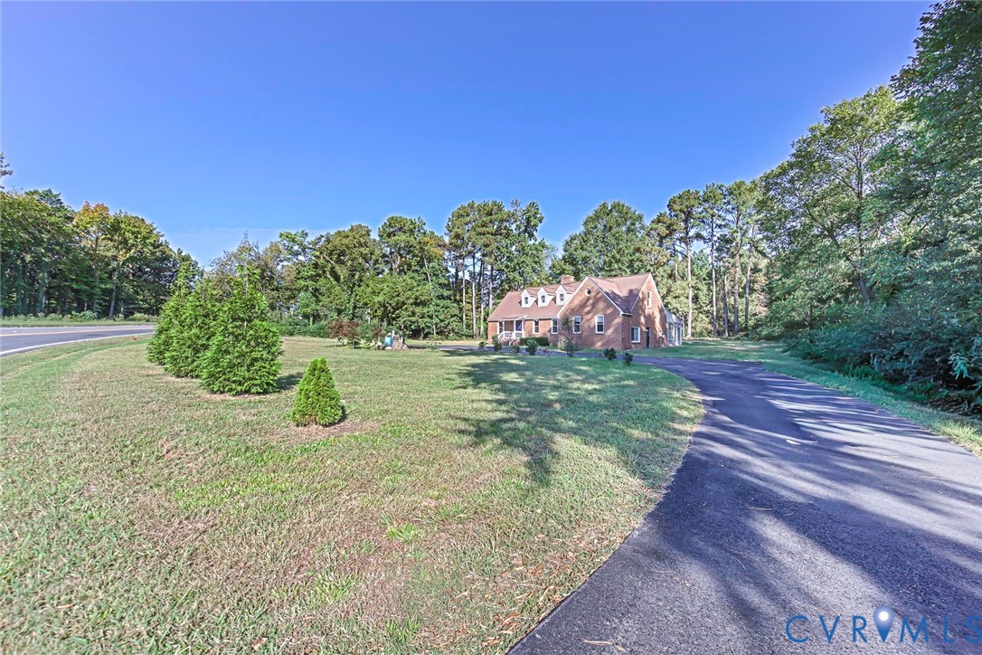 Brick exterior residence featuring a gabled roofline with dormer windows, expansive lawn, paved driveway, and mature perimeter trees Brick exterior residence featuring a gabled roofline with dormer windows, expansive lawn, paved driveway, and mature perimeter trees