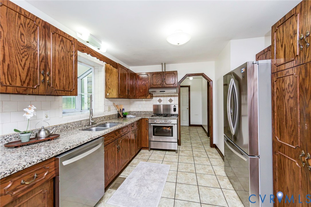 Kitchen featuring stained wood cabinetry, stone-surface countertops, stainless steel appliances, ceramic tile backsplash, and tile flooring