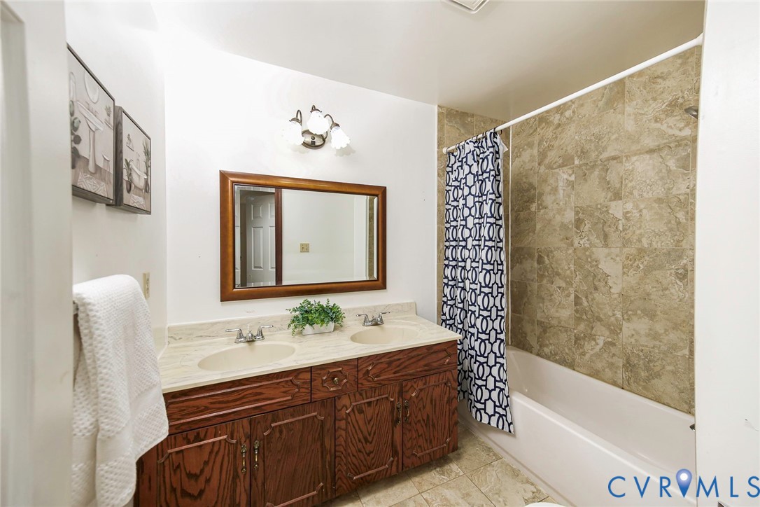 Bathroom featuring a dual-sink vanity with solid-surface countertops, wood-finish cabinetry, a framed wall mirror, and a tile-surround bathtub