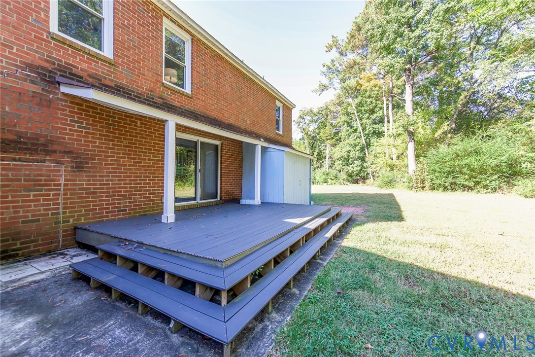 Multi-level rear deck featuring wood-finish decking, structural support columns, a sliding glass door, and exterior brick cladding, set against a tree-lined backyard
