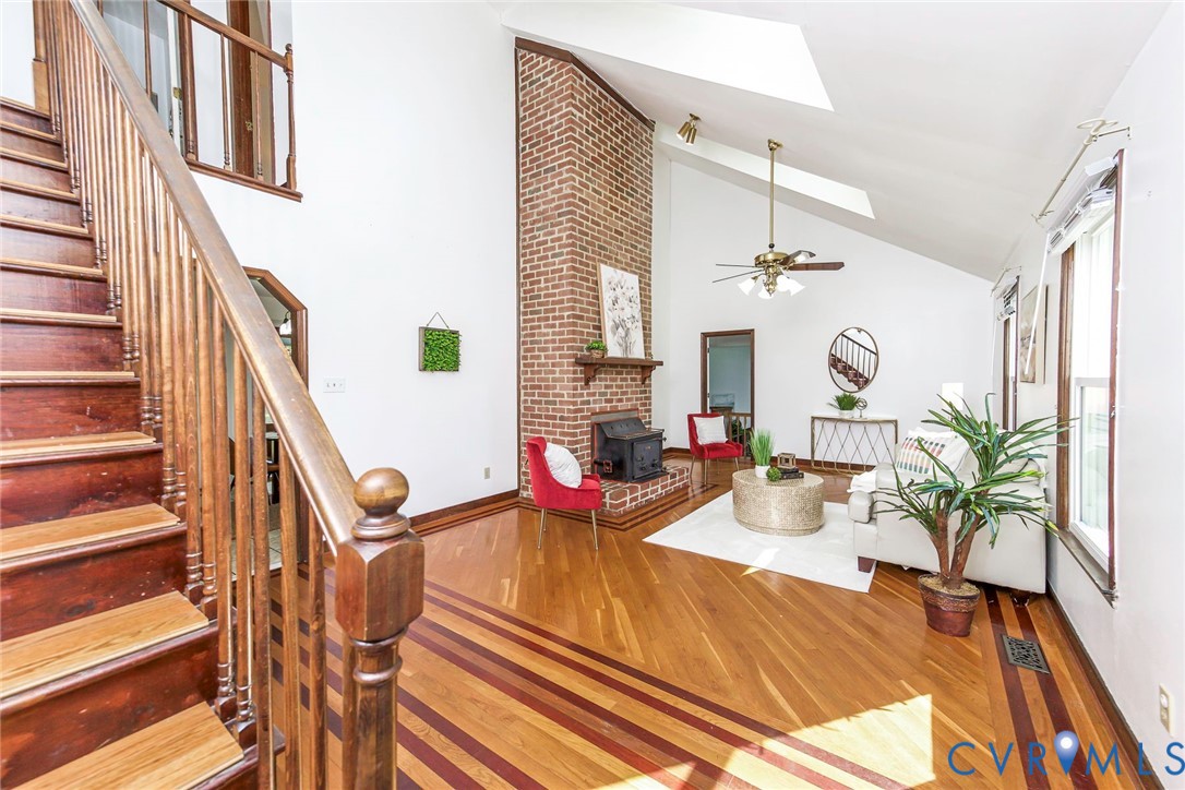 Living area featuring a floor-to-ceiling brick fireplace, wood-finish flooring with inlay detailing, vaulted ceiling with skylights, and an open staircase with a wooden balustrade Living area featuring a floor-to-ceiling brick fireplace, wood-finish flooring with inlay detailing, vaulted ceiling with skylights, and an open staircase with a wooden balustrade