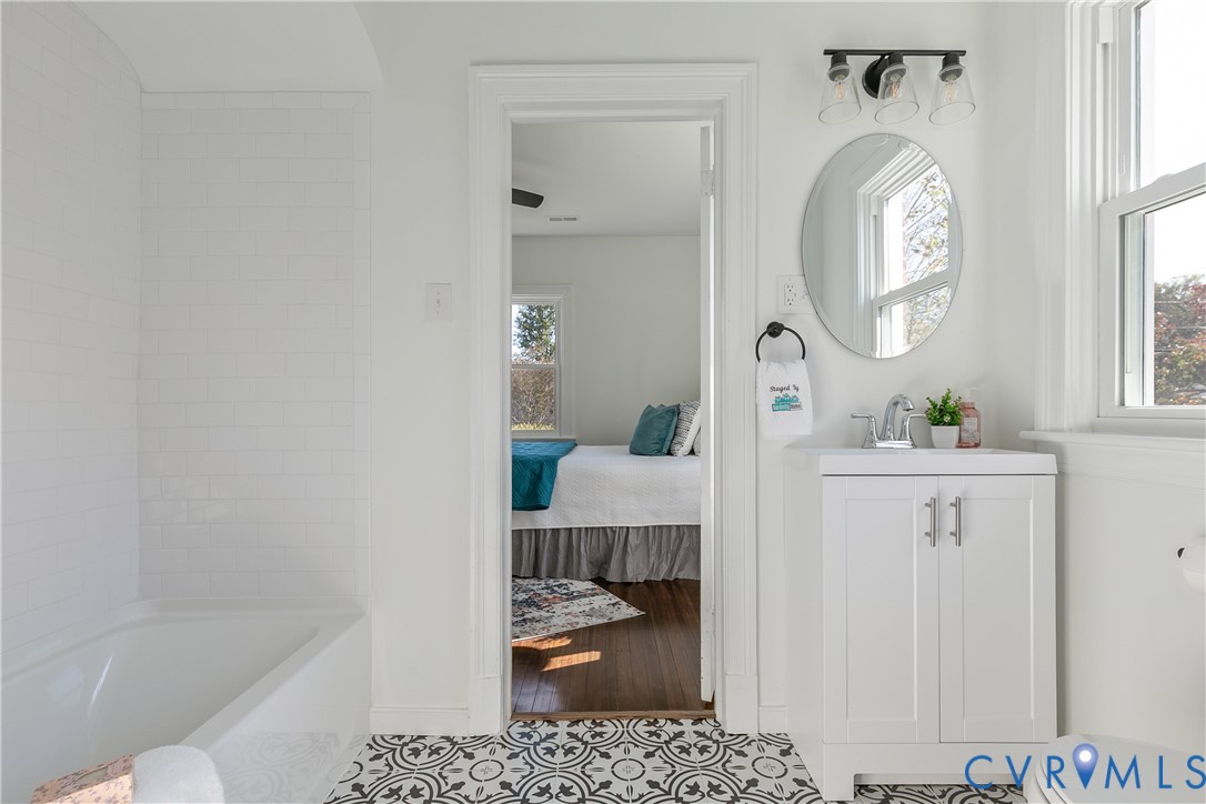 Bathroom featuring a white vanity cabinet with a stone-top sink, oval wall-mounted mirror, three-light fixture, tub-shower combination with white subway tile surround, and decorative patterned floor tile