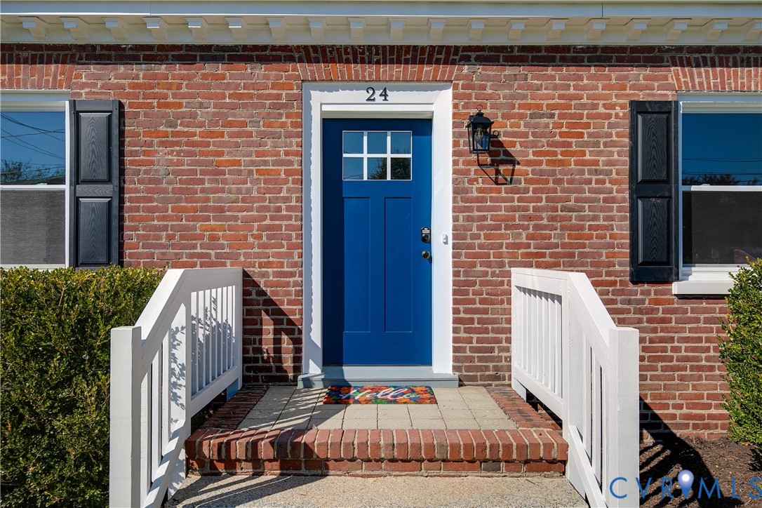 Red brick exterior featuring a blue front door with a six-pane window insert, decorative black shutters, white railings, and a brick-bordered stone stoop