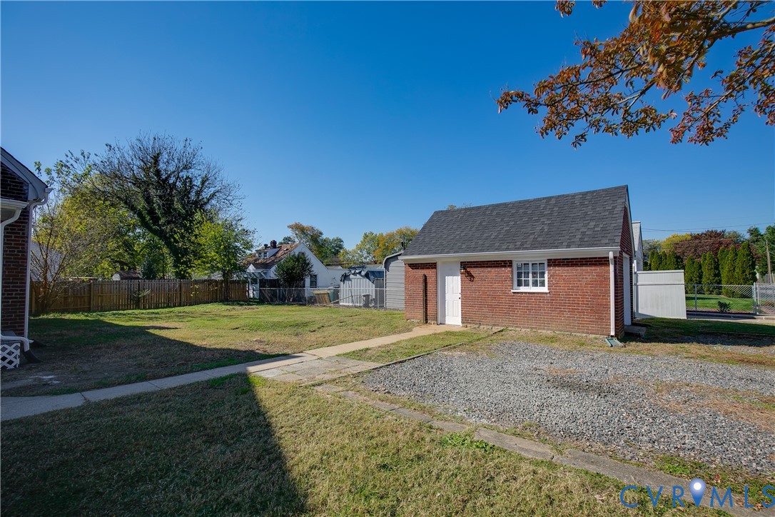 Detached brick structure featuring a gable roof, exterior lighting, and a gravel parking area, complemented by a grass yard and wooden privacy fencing