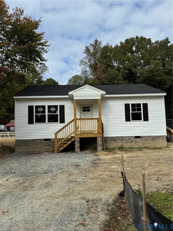 Single-story exterior featuring white vinyl siding, dark shutters, a brick foundation, a black shingled roof, and a wood-post covered entryway