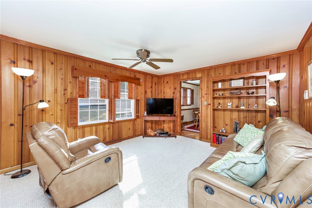 Living area featuring vertical wood-paneled walls, built-in floor-to-ceiling shelving, carpeted flooring, and a central ceiling fan