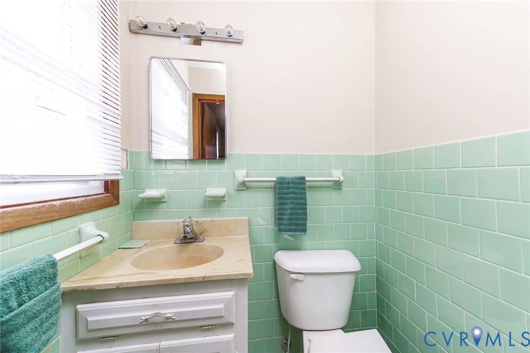 Bathroom featuring light green ceramic wall tile, a stone-finish vanity countertop with an integrated oval sink, a wall-mounted vanity mirror, and an overhead four-bulb light fixture