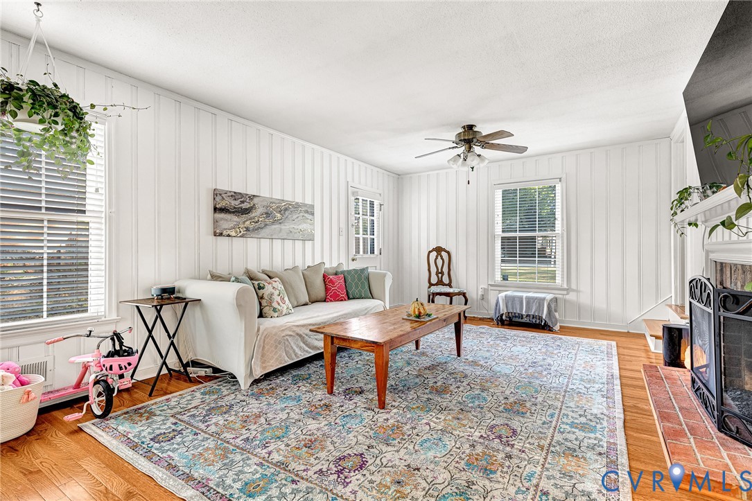 Vertical wood paneling, wood-finish flooring, brick-surround fireplace, and ceiling fan with integrated lighting
