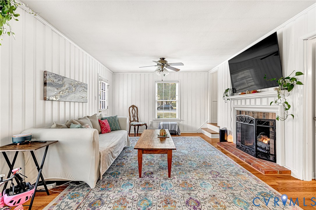 Living area featuring vertical plank wall paneling, wood-finish flooring, a wood-burning fireplace with a brick hearth, and a ceiling fan