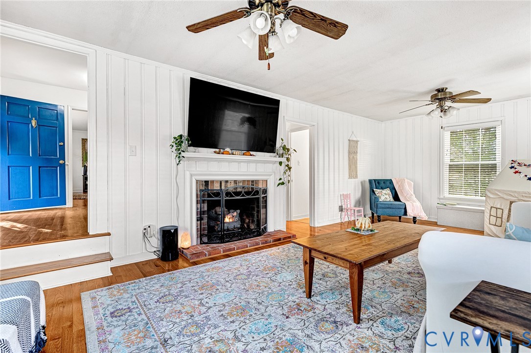Living area featuring vertical plank wall paneling, a fireplace with a brick hearth, wood-finish flooring, and dual ceiling fans