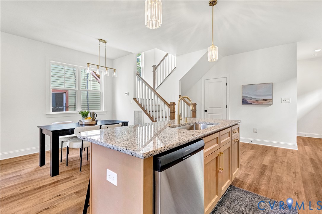 Kitchen island featuring stone countertops, a stainless steel dishwasher, and a faucet, complemented by wood-finish flooring, two pendant lights, and a wooden staircase with balusters