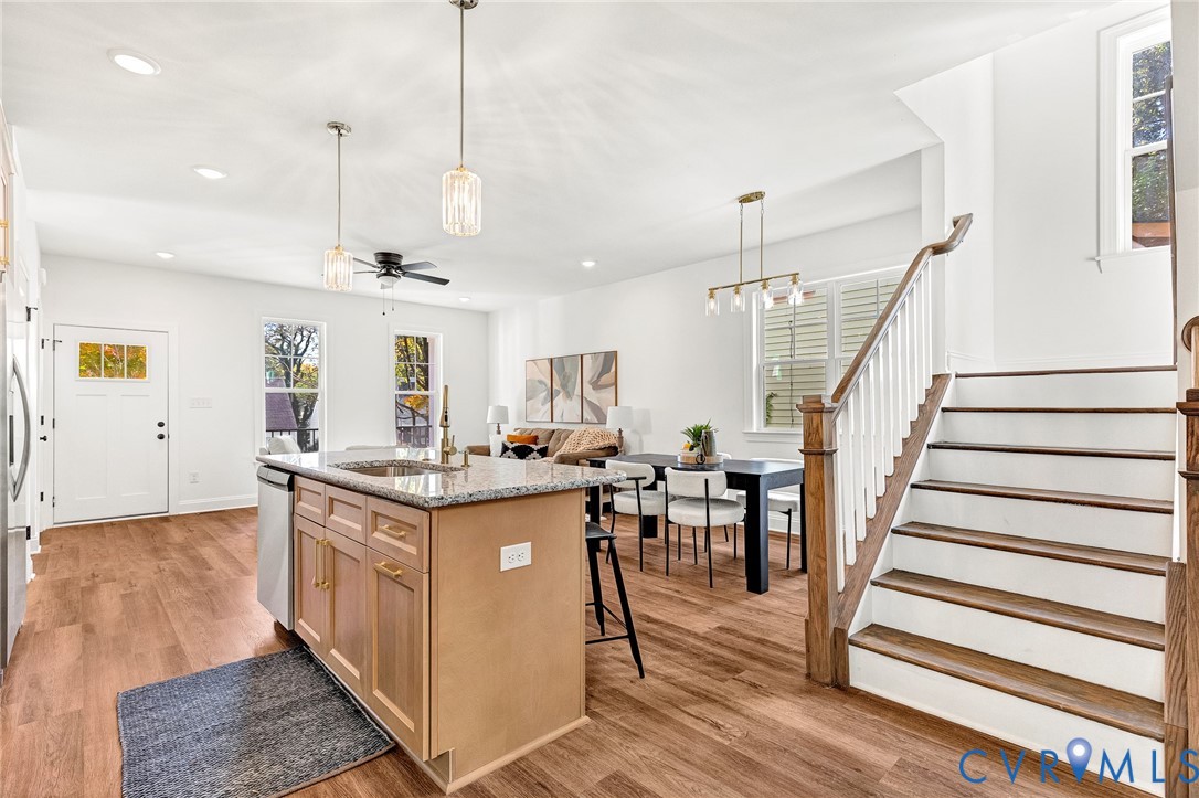 Open-concept kitchen featuring a stone-topped island with shaker-style cabinetry, wood-finish flooring, recessed lighting, and a wooden staircase with white balusters