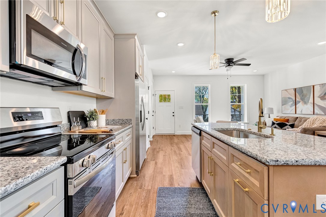 Kitchen featuring stainless steel appliances, speckled stone countertops, wood-finish flooring, shaker-style cabinetry with brass-tone hardware, and an island with an undermount sink
