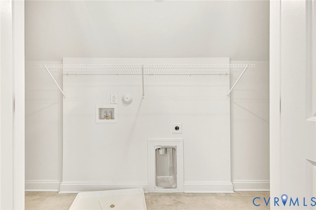 Utility area featuring a wall-mounted wire shelving unit, recessed water connection box, drainage basin, and dedicated electrical outlet
