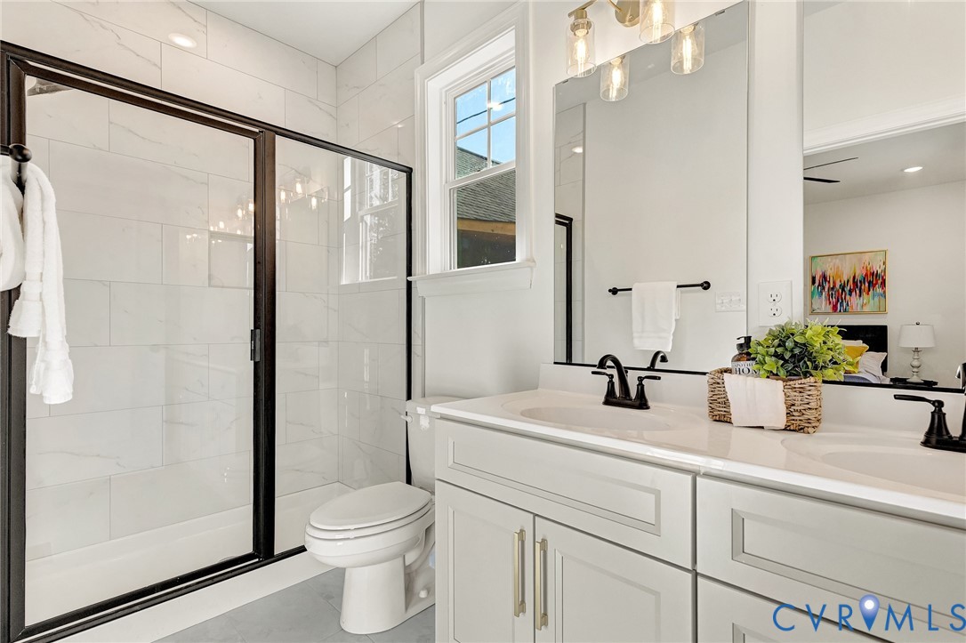 Double vanity with stone-composite countertop, light-toned shaker cabinetry, matte black hardware, and a walk-in shower featuring glass doors with black framing and light-toned tile surround