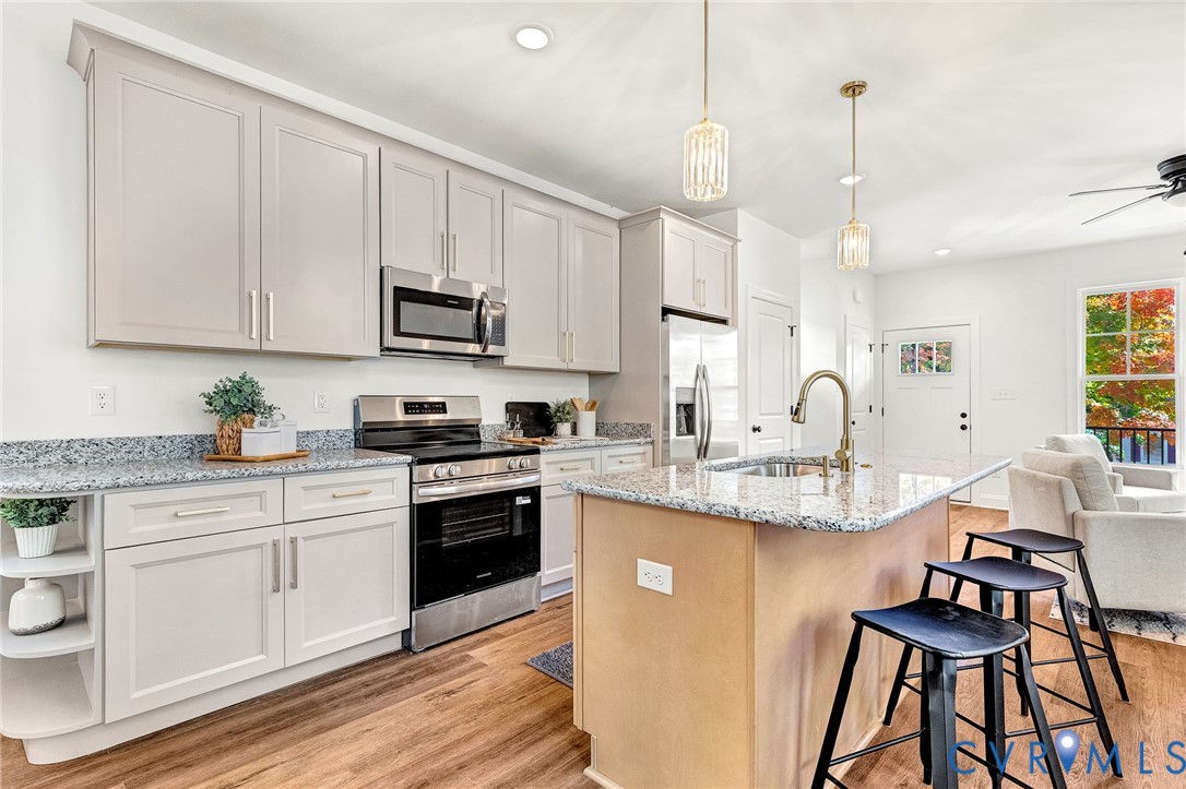 Kitchen featuring light-toned cabinetry, stone-surface countertops, stainless steel appliances, a central island with a sink, and wood-finish flooring