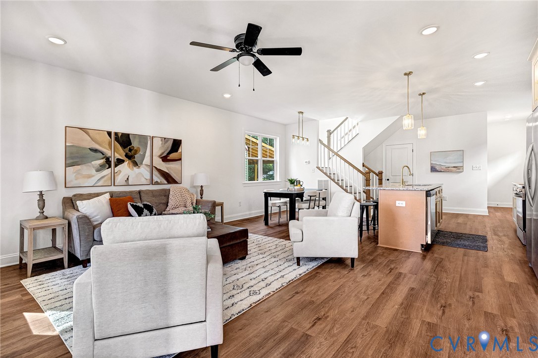 Open-concept interior featuring wood-finish flooring, recessed lighting, a ceiling fan, a kitchen island with a stone-finish countertop, and a staircase with a wooden handrail
