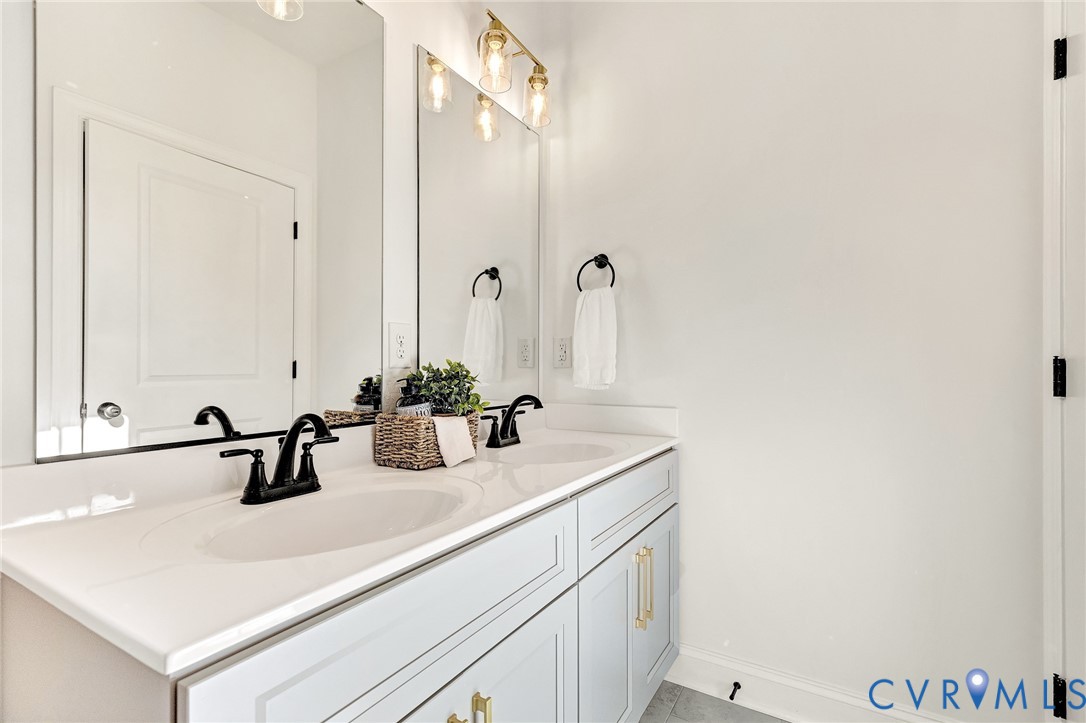 Double vanity featuring shaker-style cabinetry, white countertop with dual integrated sinks, matte black widespread faucets, and large-format wall mirrors with gold-tone overhead lighting