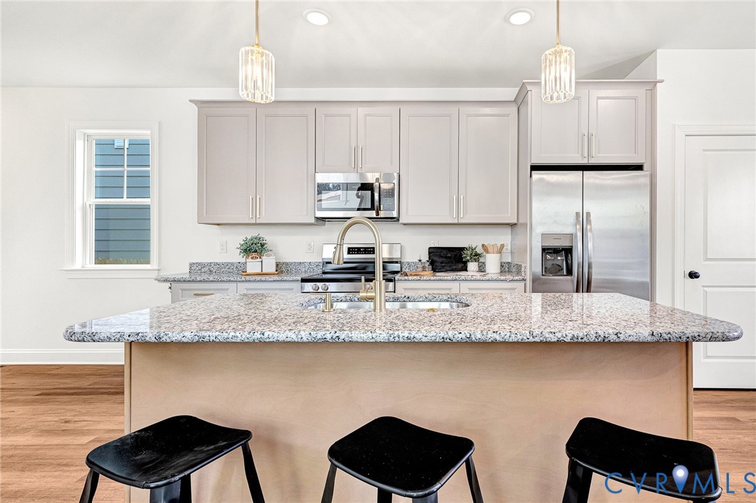 Kitchen featuring stone countertops, central island with an undermount sink, stainless steel appliances, light-toned cabinetry, and decorative pendant lighting