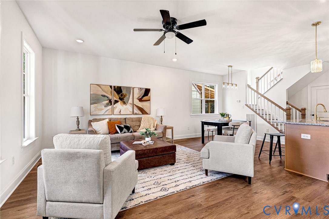 Open-concept living space featuring wood-finish flooring, a central ceiling fan, multi-pendant lighting, and a staircase with a wooden handrail