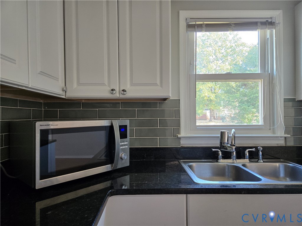 White shaker-style cabinetry, dark stone countertops, charcoal subway tile backsplash, stainless steel double-basin sink with pull-down faucet, and built-in stainless steel microwave