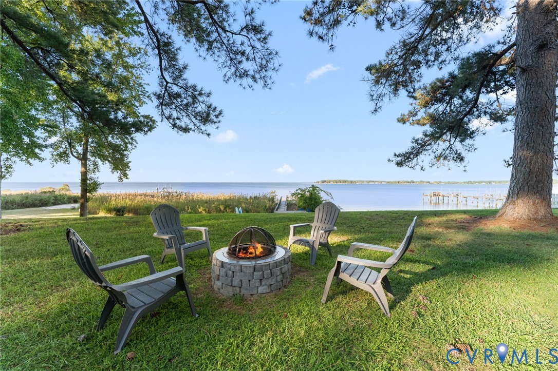 Waterfront grounds featuring a circular stone fire pit, sprawling lawn, mature trees, and a wooden dock