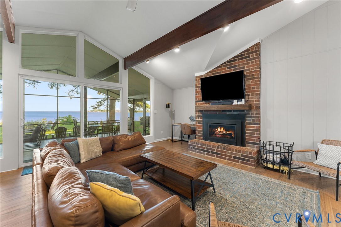 Vaulted ceilings with exposed wood beams, full-height brick fireplace, wood-finish flooring, and expansive windows featuring water scenery
