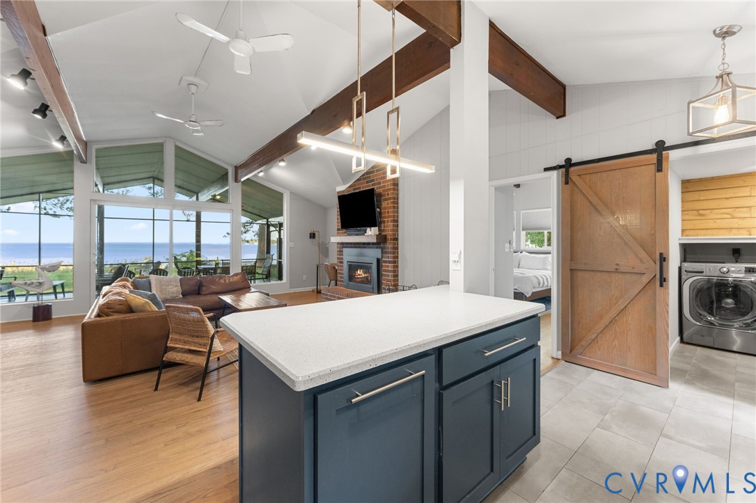 Vaulted ceiling with exposed wood beams, brick fireplace, expansive floor-to-ceiling windows, center island with stone-finish countertop, and sliding barn door