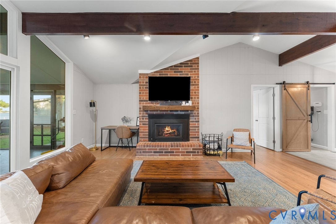 Vaulted ceiling with exposed wooden support beams, brick fireplace featuring a wood mantel, floor-to-ceiling windows, and interior sliding wooden barn door