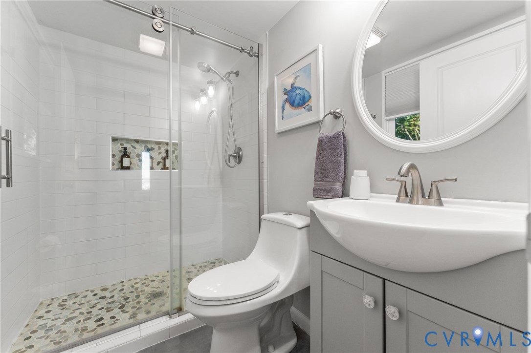 Bathroom featuring a walk-in shower with a sliding glass door, white subway tile surround, integrated stone-tiled niche, pebble-stone flooring, and a grey vanity with a rounded basin and oval mirror