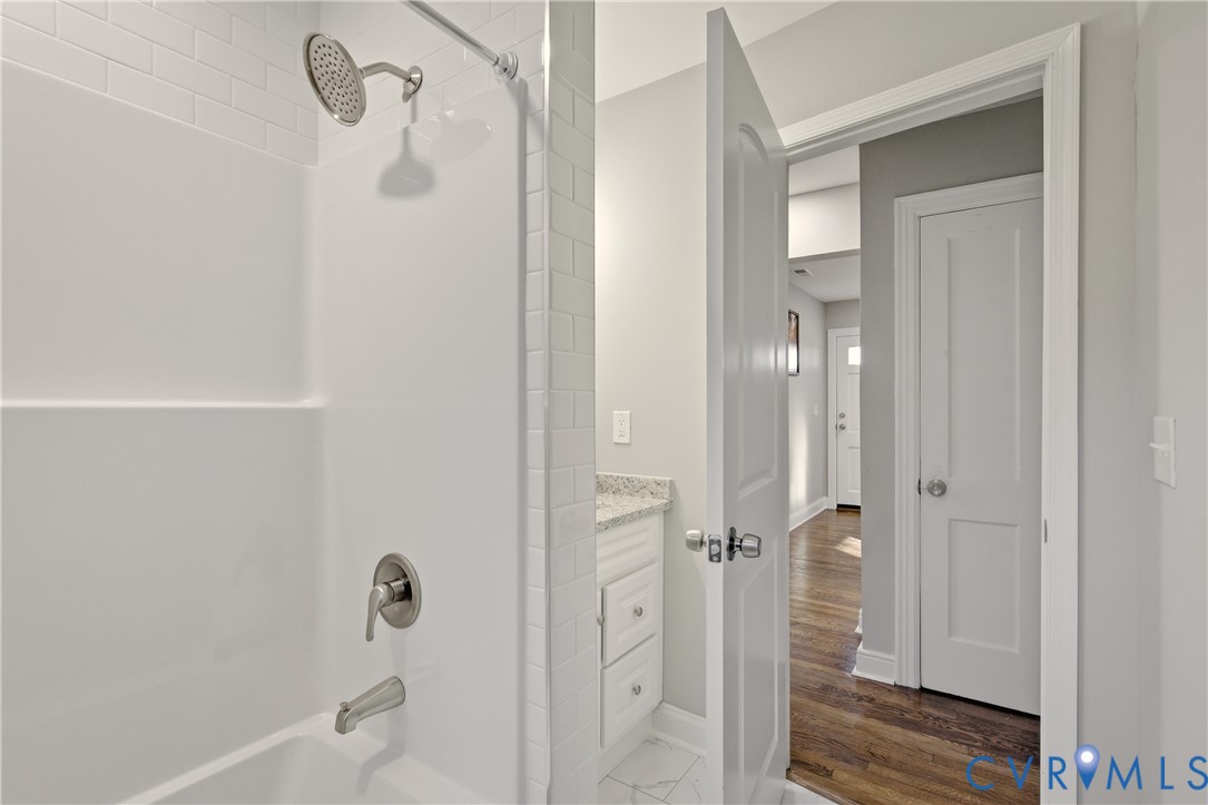 Bathtub with a shower head, subway-tile surround, stone-finish vanity countertop, and wood-finish flooring
