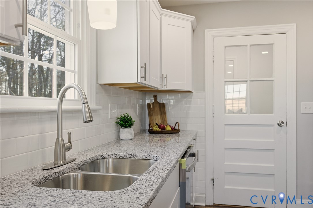 Kitchen featuring white shaker-style cabinetry, speckled stone-finish countertops, a dual-basin stainless steel sink with a high-arch faucet, white subway tile backsplash, and a multi-pane glass door