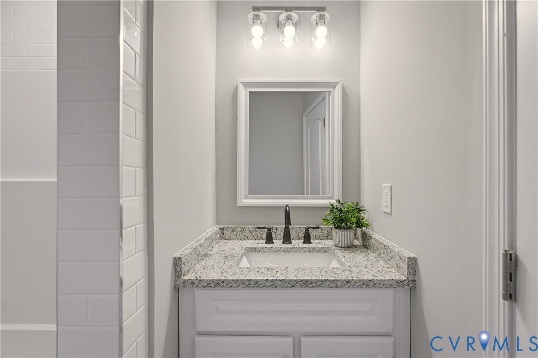 Bathroom vanity featuring a stone-composite countertop, undermount sink, three-light fixture, framed mirror, and white shaker-style cabinetry