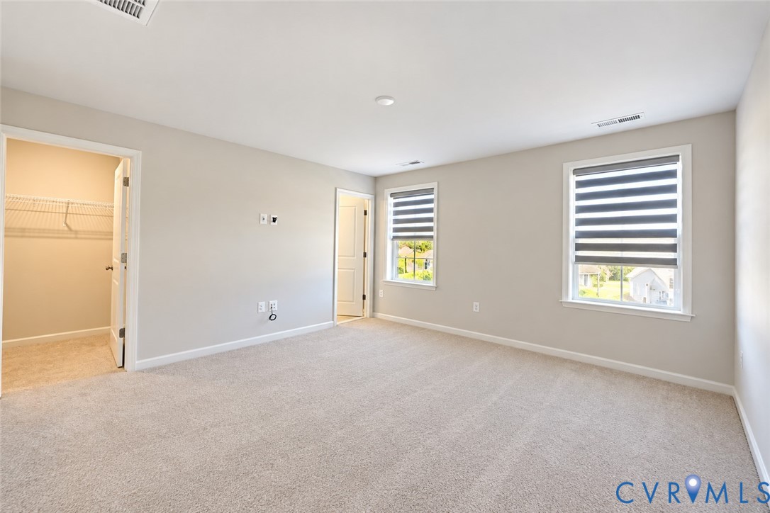 Neutral-toned room featuring plush wall-to-wall carpeting, dual windows with banded roller shades, and a walk-in closet with built-in wire shelving