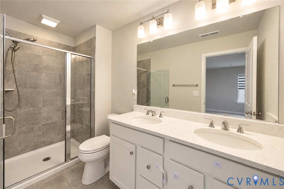 Bathroom featuring a dual-sink vanity with a solid-surface countertop, full-width mirror, glass-enclosed shower with tiled surround, and multi-light fixtures