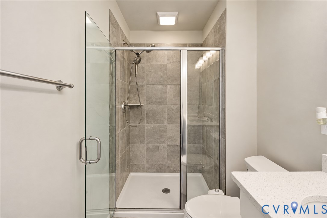 Walk-in shower featuring a glass enclosure, oversized neutral tile surround, and adjustable handheld showerhead, complemented by a stone-composite countertop with a recessed sink