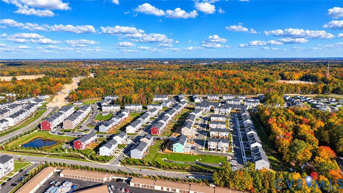 Aerial view of residential community featuring multi-unit buildings, paved streets, detention pond, and surrounding wooded area